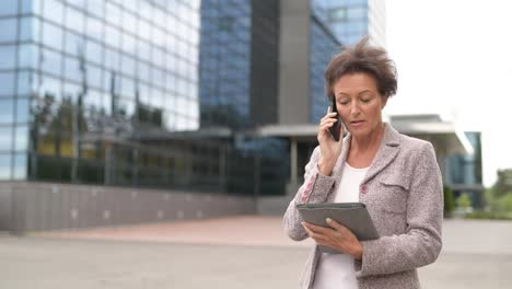 Feliz-mujer-de-negocios-madura-usando-el-teléfono-y-la-tableta-digital-en-la-ciudad-al-aire-libre