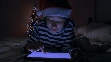 Mittlerer-Schuss.-Asiatische-von-kleinen-Mädchen-mit-digitalen-Tablet,-während-auf-dem-Bett-im-Schlafzimmer-mit-Weihnachtsbaum-in-der-Nacht-dekoriert-liegen