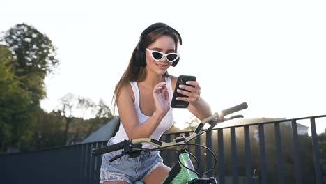 Close-up-of-smiling-young-woman-in-headphones-sitting-on-the-bike-and-listening-music