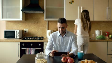 Joven-feliz-con-tableta-digital-sentado-en-la-cocina-mientras-su-novia-cocina-en-casa