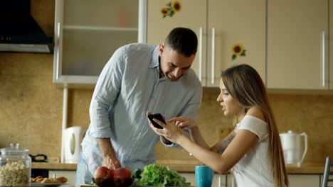 Attraktives-Paar-in-der-Küche-am-frühen-Morgen.-Schöne-Mädchen,-die-gemeinsame-Nutzung-von-social-Media-auf-Smartphone-mit-ihrem-Freund