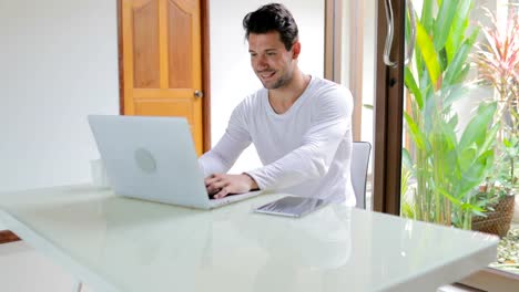 Young-Man-Working-With-Laptop-Computer-Sitting-Table-At-Home-Smiling-Guy-Typing