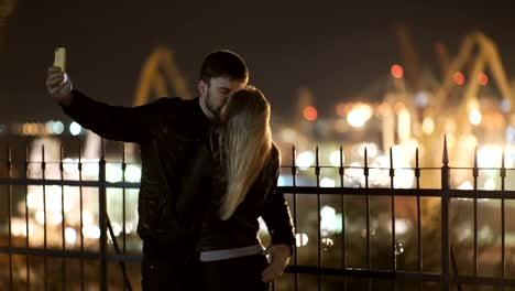 Young-happy-couple-take-selfie-photos-at-camera-on-smart-mobile-cell-phone.-Night-lights-of-port