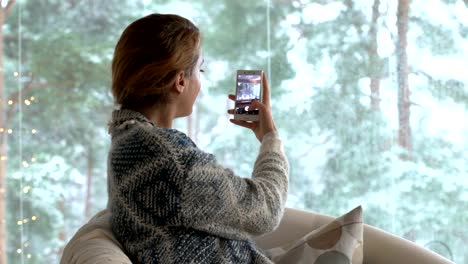 Eine-Frau-benutzt-eine-Smartphone-in-ein-gemütliches-Wohnzimmer-mit-dem-großen-schneebedeckten-Fenster-sitzen