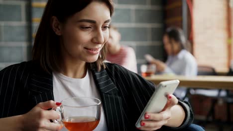 Lächelnd-Unternehmerin-Surfen-Smarpthone-und-Tee-zu-trinken,-während-haben-Bruch-in-der-geschäftigen-Büro