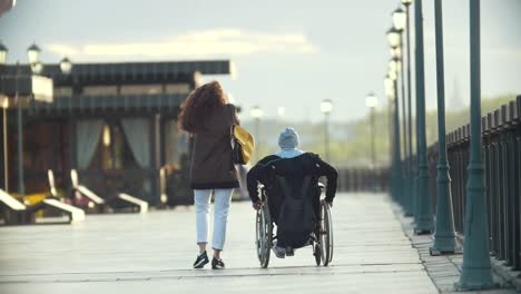 Mujer-joven-feliz-con-un-hombre-discapacitado-en-silla-de-ruedas-caminar-juntos-en-el-muelle