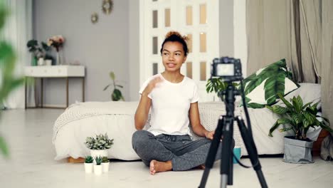 Chica-afro-americano-apuesto-blogger-creativo-está-grabando-video-sobre-plantas-sentado-en-el-piso-de-su-apartamento-y-habla-mirando-a-cámara-en-el-trípode.