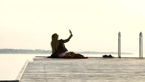 Una-hermosa-niña-sentada-en-un-muelle-en-la-playa-tomando-un-instagram-storry-selfie,-manteniendo-el-signo-de-la-paz-en-la-puesta-de-sol