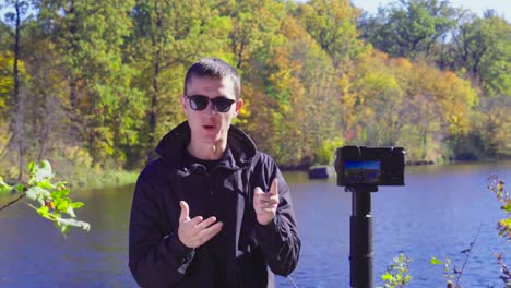 Brunette-man-blogger-in-sunglasses-on-river-bank-is-recording-video-for-vlog-using-camera