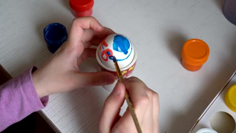 The-child-is-preparing-for-the-holiday-and-draws-on-the-Easter-egg-symbols-of-blue-and-red-paint,-sitting-at-the-table.-Close-up.-The-view-from-the-top.-4K.