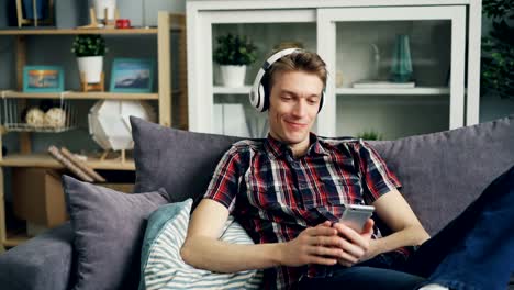 El-joven-feliz-en-los-auriculares-está-escuchando-música-y-usando-el-teléfono-inteligente-en-casa