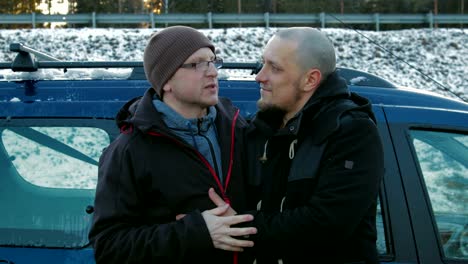 Two-LGBT-guys,-bald-with-a-beard-and-wearing-glasses-and-a-hat-hugging-and-chatting-on-the-side-of-the-road-near-the-car