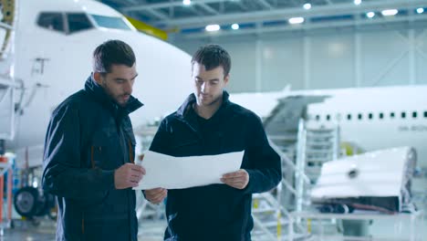 Team-of-Aircraft-Mechanics-and-Engineers-Waking-Through-Airplane-Maintenance,-Design-and-Development-Facility.-Professionals-Consulting-Blueprints-and-Talking