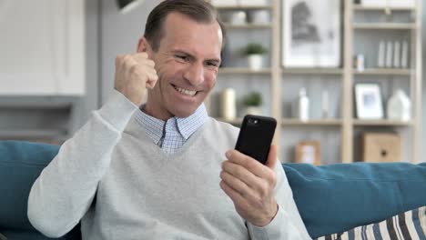 Mittelalter-Mann-Cheering-auf-Smartphone-beim-Sitzen-auf-Couch