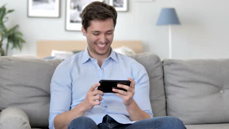 Hübsche-Young-Man-Cheering-Erfolg-auf-Smartphone-beim-Sitzen-auf-Couch