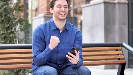 Young-Man-Celebrating-Win-on-Smartphone,-Sitting-Outdoor-on-Bench