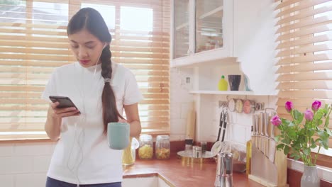 Young-Asian-teenager-woman-drinking-warm-coffee-using-smartphone-listening-music-and-checking-social-in-kitchen-at-home.-Lifestyle-woman-relax-in-morning-at-home-concept.