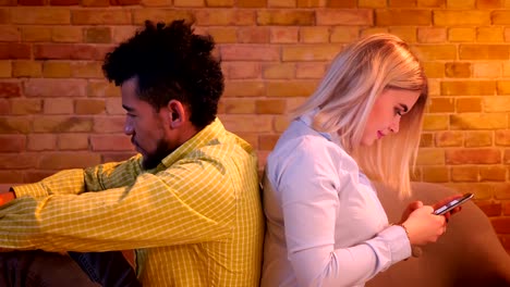 Close-up-shot-of-african-guy-and-caucasian-girl-sitting-back-to-back-on-sofa-watching-into-their-smartphones-at-home.