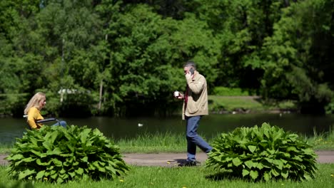 Lockdown-side-view-shot-of-active-senior-woman-in-wheelchair-riding-down-path-in-green-park,-mature-man-with-takeaway-coffee-cup-passing-by-her-talking-on-cell-phone