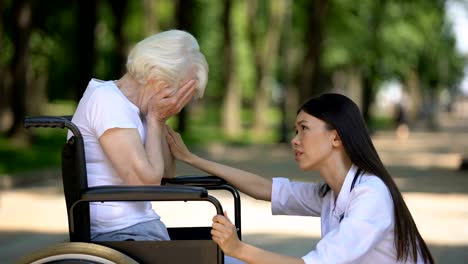 Female-volunteer-supporting-sad-elderly-woman-in-wheelchair-in-hospital-park