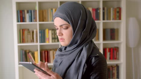 Nahaufnahme-Porträt-von-jungen-attraktiven-muslimischen-Studentin-in-Hijab-mit-dem-Tablet-lächelnd-Blick-auf-die-Kamera-in-der-College-Bibliothek-in-Innenräumen