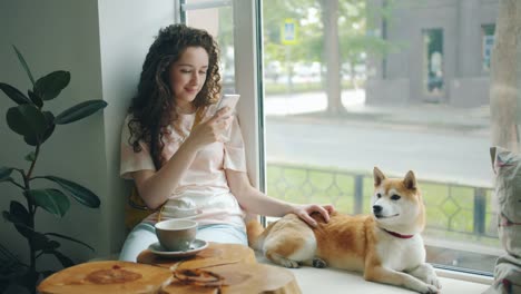 Attractive-woman-using-smartphone-sitting-on-window-sill-in-cafe-with-puppy