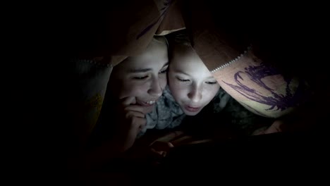 Two-smiling-little-girls-watching-pictures-on-a-tablet,-lying-at-night-under-a-blanket-on-the-bed-in-the-dark.-Concept-video.-Internet-addiction.-Close---up-of-children's-faces.-Raw-video.-4K.