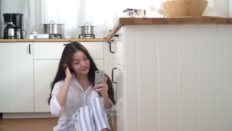 4K-Medium-Dolly-Schuss-von-jungen-asiatischen-Frau-sitzt-auf-Küchenboden-zu-Hause-mit-Smartphone-mit-drahtloser-Technologie-für-Social-Media-Kommunikation.-Asiatische-Teenager-Mädchen-genießen-auf-Küchenboden-mit-Smartphone.