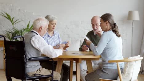 Grupo-de-cuatro-personas-mayores-jubiladas,-dos-hombres-y-dos-mujeres,-divirtiéndose-sentados-a-la-mesa-y-jugando-a-las-cartas-juntos-en-la-sala-común-de-residencia-de-ancianos,-rastreando-la-toma