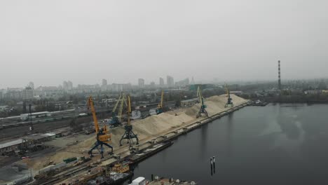 Luftaufnahme-der-Industriestadt-in-Smog-und-Nebel-mit-Baukränen,-die-an-Docks-arbeiten.-Sandkahn-und-Lastwagen-transportieren-Sand.