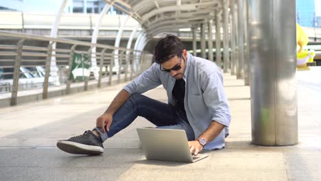 young-businessman-in-smart-casual-wear-sitting-on-street-using-laptop-and-typing-a-message-in-urban-city-outdoors.-freelancer-working-on-floor-outside-Chatting-to-customer.-blue-jeans