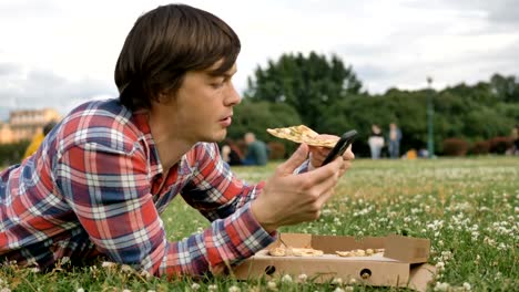 man-lying-on-the-grass-eating-pizza-and-using-a-smartphone-in-a-city-park-on-nature