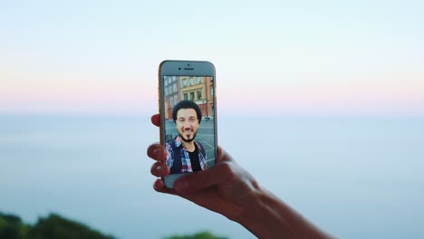 Mano-sosteniendo-el-teléfono-inteligente-durante-la-videollamada-con-el-hombre-en-frente-del-mar