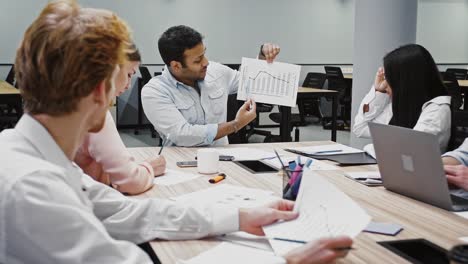 Joven-jefe-confiado-discutiendo-detalles-de-la-nueva-startup-con-diversos-colegas,-sentados-en-la-mesa-en-la-oficina-moderna