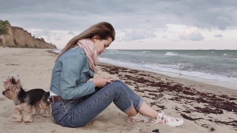 Young-beautiful-girl-is-writing-a-message-on-a-mobile-phone-by-the-sea.-A-small-dog-is-walking-nearby