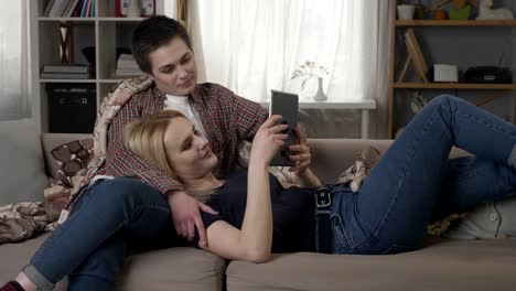 Lesbian-couple-is-resting-on-the-couch,-using-tablet-computer,-scrolling-photos-on-tablet,-holding-hands,-smiling-60-fps