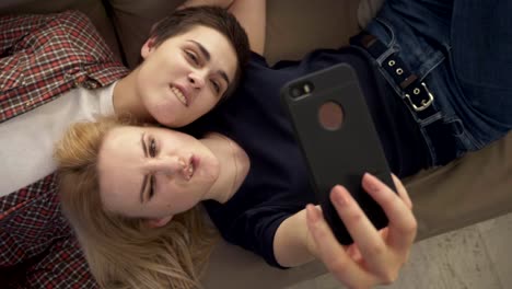 Two-young-lesbian-girls-lie-on-the-couch,-do-selfie-on-a-smartphone,-lovers,-lgbt,-young-couple,-teenagers-top-shot-60-fps