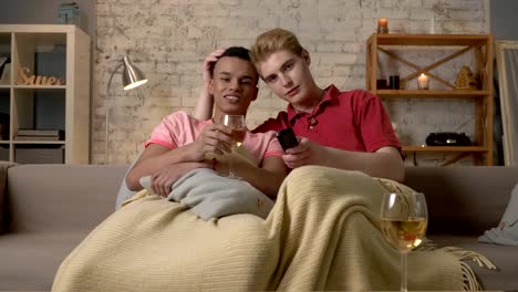 Multinational-gay-couple-sitting-on-couch-covered-with-a-warm-blanket,-watch-TV,-use-the-remote-control,-look-at-the-camera.-Homeliness,-romantic-evening,-cuddles,-hugs,-happy-LGBT-family-concept.-60-fps