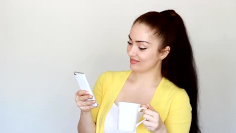 Portrait-of-a-beautiful-brunette-girl-who-drinks-a-hot-drink-from-a-cup-and-holds-a-mobile-phone-in-his-hand.-Young-woman-reads-social-network-and-looks-internet-on-smartphone-screen-and-smiles