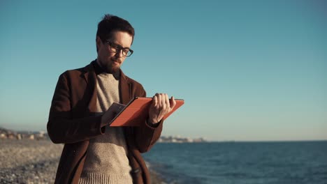 Bearded-man-is-using-flatbed-computer-sitting-on-seashore-with-panorama-of-city