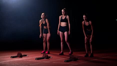 Tres-chicas-deportivas-en-ropa-de-color-negro-del-atleta-en-la-noche-en-la-cinta-se-iniciará-para-la-carrera-en-la-distancia-sprint-desde-la-posición-sentada