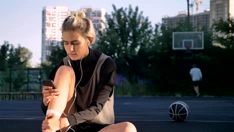 Wunderschöne-blonde-Frau-Musik-über-Kopfhörer-hören-und-tippen-auf-Telefon,-sitzt-auf-Platz,-Mann-im-Hintergrund-wirft-ball