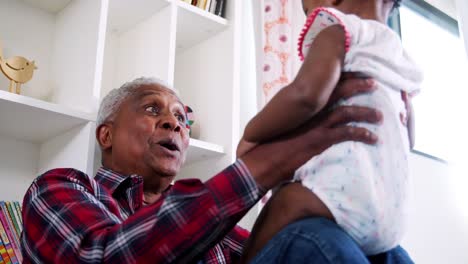 Grandfather-sitting-on-sofa-at-home-lifting-baby-granddaughter-into-air-and-smiling---shot-in-slow-motion