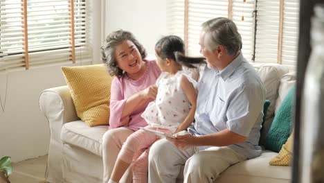 Little-girl-comes-to-hug-her-grandfather-and-grandfather-while-they-playing-tablet.-Kid-playing-with-senior-man-and-woman-with-happy-emotion.-people-with-family,-lifestyle,-technology-concept.