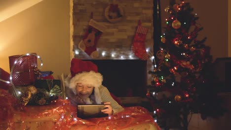 Glückliches-kleines-Mädchen-mit-Tablet-auf-einen-Videoanruf-zu-Weihnachten-in-eingerichteten-Wohnzimmer-mit-Baum-und-Schornstein.