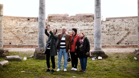 Smiling-young-European-girl-and-happy-senior-tourist-group-taking-selfie-near-old-amphitheater-ruins-in-Ostia,-Italy.