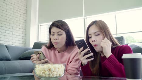 Asian-women-using-smartphone-and-eating-popcorn-in-living-room-at-home,-group-of-roommate-friend-enjoy-funny-moment-while-lying-on-the-sofa.-Lifestyle-women-relax-at-home-concept.