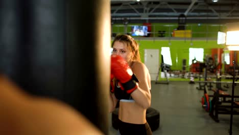 Young-woman-boxer-hit-punching-bag-during-pre-match-warm-up-with-her-trainer-in-boxing-club