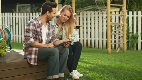 Young-Man-And-Woman-Using-Touchscreen-Tablet-Computer-Together,-they-are-on-Backyard-at-Bright-Summer-Day.