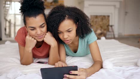 Zwei-fröhliche-gemischte-Rasse-lustige-Freundinnen-teilen-Soziale-Medien-mit-Tablet-Computer-und-Gespräche-liegen-im-Bett-zu-Hause